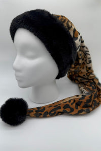A long leopard-print plush hat with a wide black faux fur trim and a large black faux fur pom at the end, displayed on a white mannequin head. The hat drapes down to the side, showcasing its extended length and bold animal-print pattern.