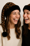 Two women wearing leopard print hats with pom-poms, laughing together.