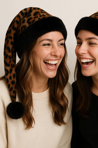 Two women wearing leopard print hats with pom-poms, laughing together.