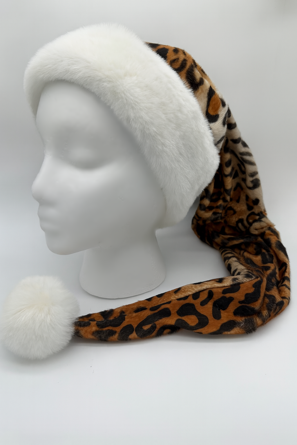 The image shows a white Styrofoam mannequin head wearing a long leopard-print Santa-style hat. The hat features a fluffy white fur trim around the brim and a matching white pom-pom at the end of the extended tail, which drapes down to the front.