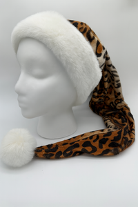 The image shows a white Styrofoam mannequin head wearing a long leopard-print Santa-style hat. The hat features a fluffy white fur trim around the brim and a matching white pom-pom at the end of the extended tail, which drapes down to the front.
