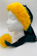 A festive hat displayed on a white mannequin head. The hat is made of deep green Velvet with a wide, fluffy yellow faux-fur trim and a large yellow pom-pom at the end. Small decorative footpom-pom-shaped buttons are attached along the green Velvet fabric.