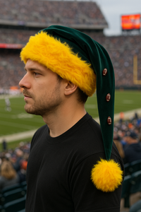 A light-skinned man with a short beard is sitting in a football stadium, facing right, wearing a green Velvet Santa-style hat with bright yellow faux fur trim, a large yellow pom-pom, and small football-shaped buttons. The hat’s long tail extends down to about 8 inches below his shoulder, and the blurred field and crowd are visible in the background.