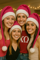 Four smiling young women at a sorority party wear matching bright pink Santa hats with white faux-fur trim and pom-poms that hang about eight inches below their shoulders. They stand close together in a warmly lit room decorated with soft string lights, creating a festive and joyful holiday atmosphere.