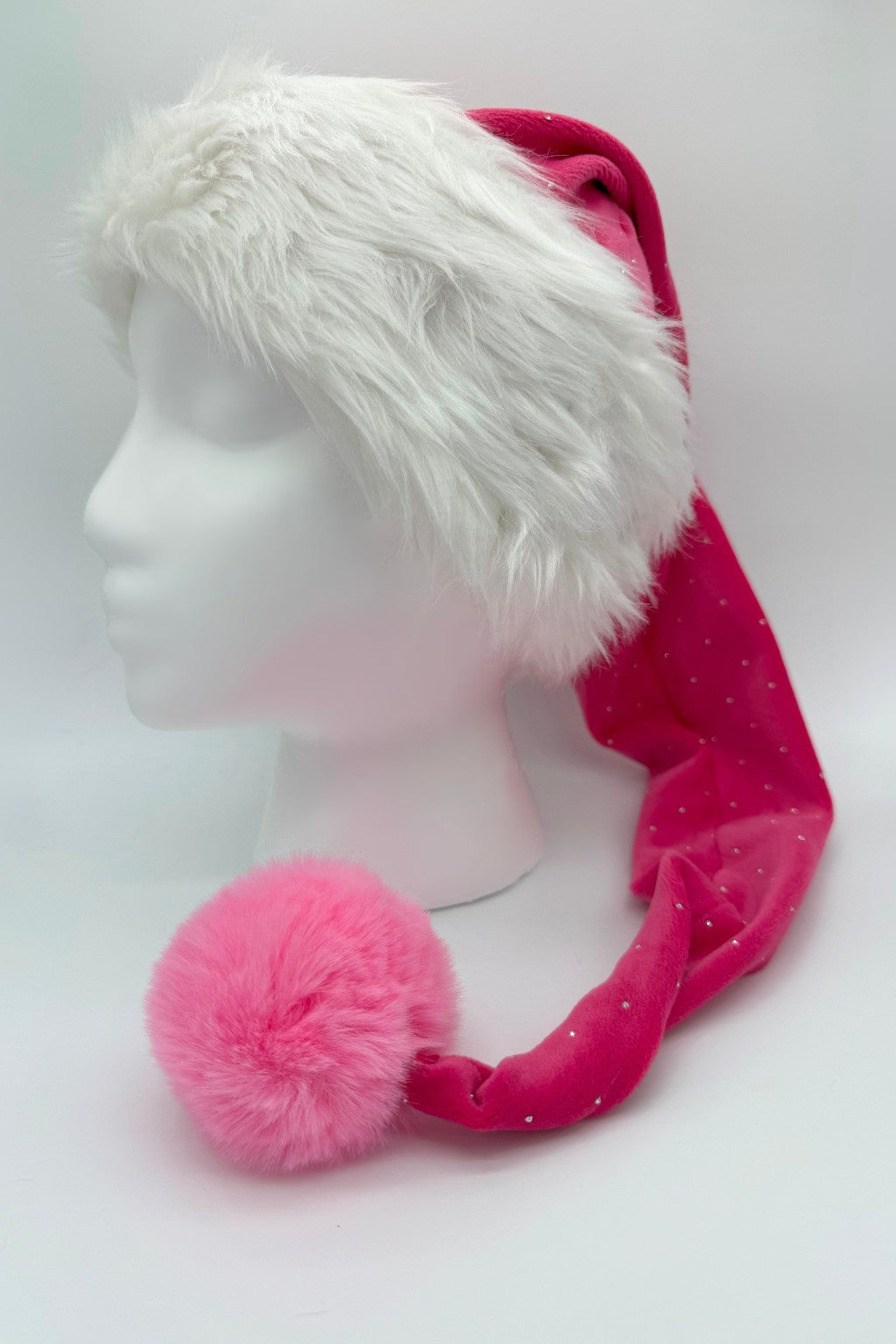 A festive Santa-style hat displayed on a white mannequin head. The hat has a fluffy white faux fur trim and a long, bright pink body with small sparkly dots. A large, fluffy pink pom-pom sits at the tip of the hat, resting near the mannequin’s shoulder.
