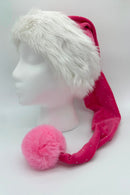 A festive Santa-style hat displayed on a white mannequin head. The hat has a fluffy white faux fur trim and a long, bright pink body with small sparkly dots. A large, fluffy pink pom-pom sits at the tip of the hat, resting near the mannequin’s shoulder.