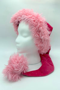 A mannequin head displays a festive hat with a fluffy pink faux fur trim and a long, pointed body made of dark pink fabric decorated with small white dots. The hat ends in a large, fluffy pink pom-pom resting near the mannequin’s shoulder.