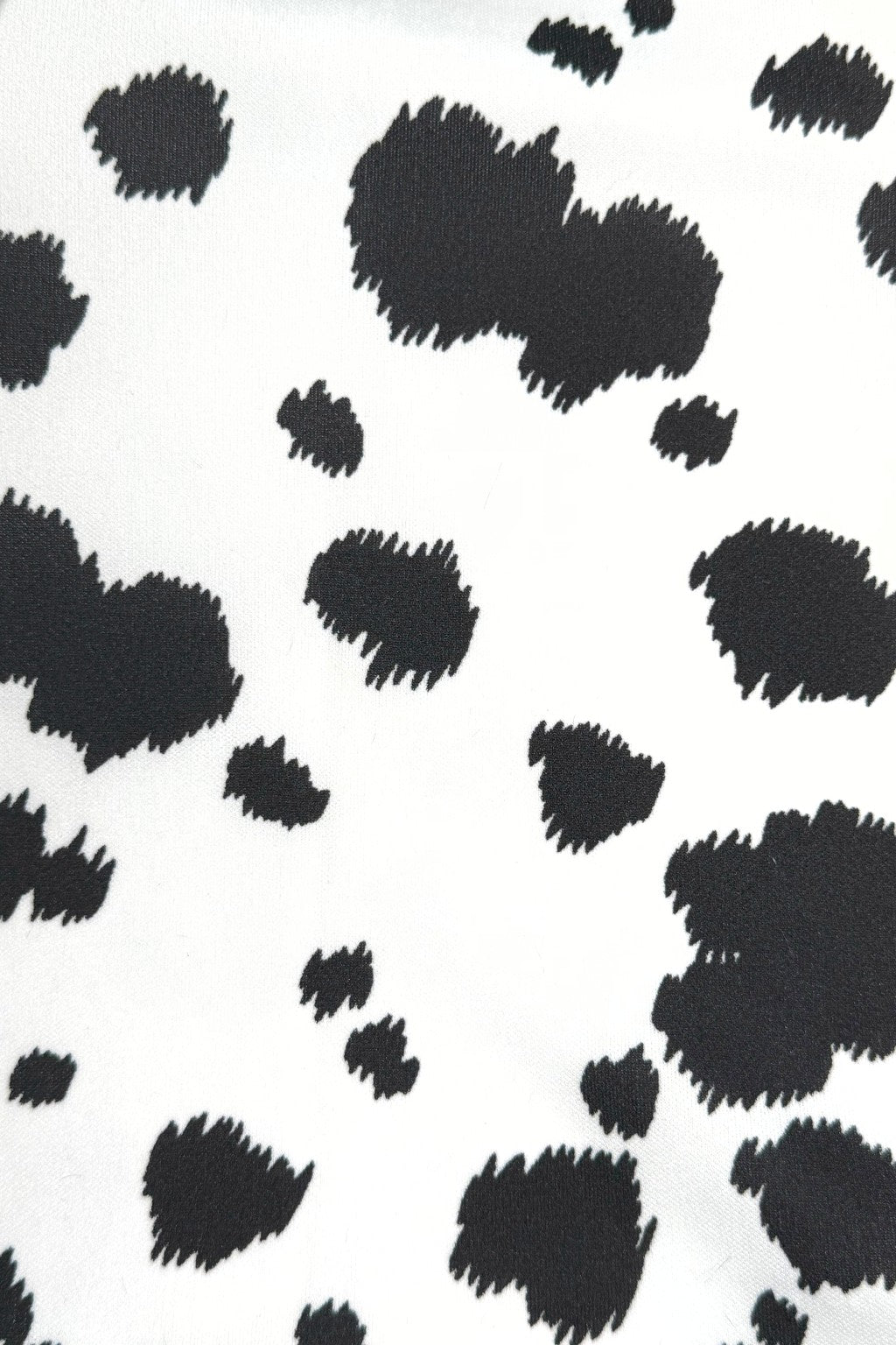 Close-up of a white fabric with an irregular black cow print pattern, featuring scattered black spots of varying sizes across the surface.