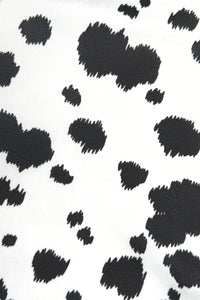 Close-up of a white fabric with an irregular black cow print pattern, featuring scattered black spots of varying sizes across the surface.