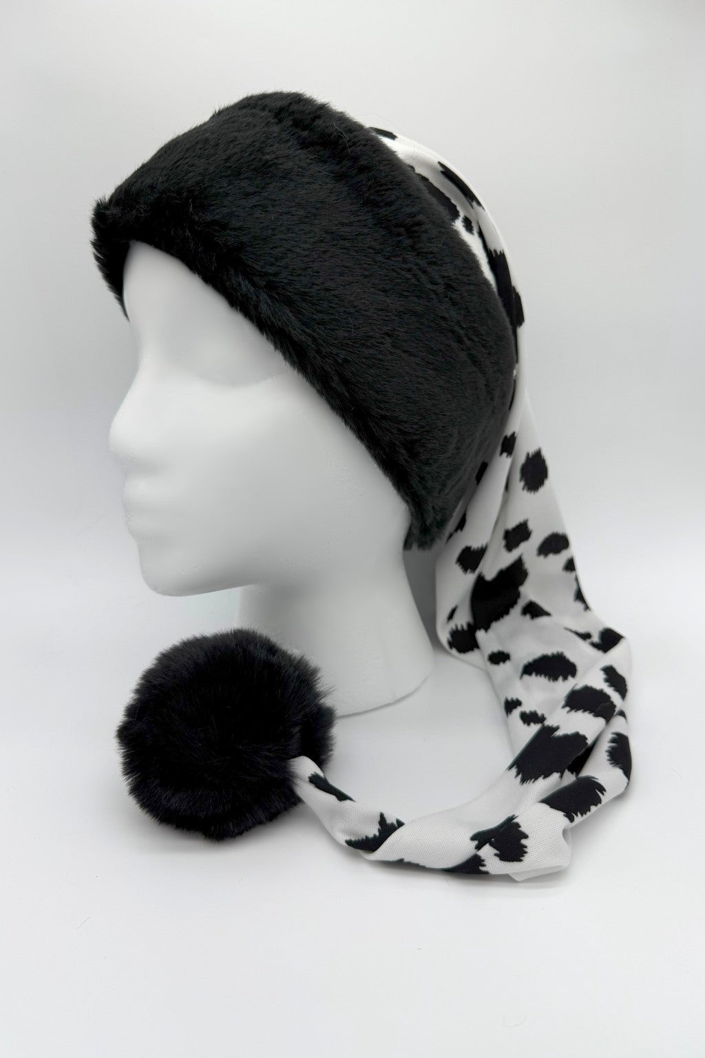 A black and white cow-print Santa-style hat displayed on a white mannequin head. The hat has a thick, plush black faux fur trim around the forehead and a long, draping tail made of silky cow-print fabric that ends with a large, fluffy black pom-pom resting on the surface. The background is plain white with reflective side panels.
