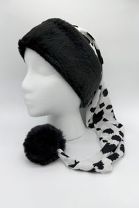 A black and white cow-print Santa-style hat displayed on a white mannequin head. The hat has a thick, plush black faux fur trim around the forehead and a long, draping tail made of silky cow-print fabric that ends with a large, fluffy black pom-pom resting on the surface. The background is plain white with reflective side panels.