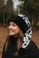 A woman outdoors in a wooded area smiles while wearing a black faux fur and cow-print Santa-style hat with a long tail ending in a black pom-pom.