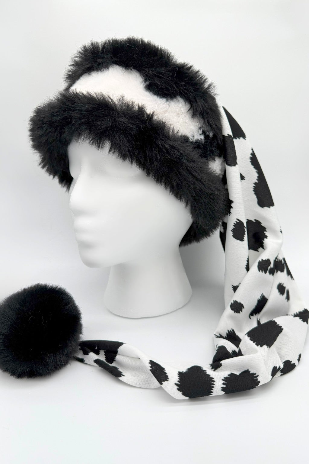 Black and white faux fur Santa-style hat displayed on a white mannequin head. The hat features a fluffy black fur brim with white fur center, and a long draping tail made of silky fabric in a bold black-and-white cow print pattern, ending with a large black fur pom-pom.
