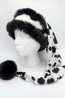 Black and white faux fur Santa-style hat displayed on a white mannequin head. The hat features a fluffy black fur brim with white fur center, and a long draping tail made of silky fabric in a bold black-and-white cow print pattern, ending with a large black fur pom-pom.