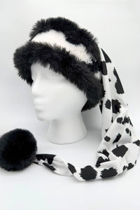 Black and white faux fur Santa-style hat displayed on a white mannequin head. The hat features a fluffy black fur brim with white fur center, and a long draping tail made of silky fabric in a bold black-and-white cow print pattern, ending with a large black fur pom-pom.