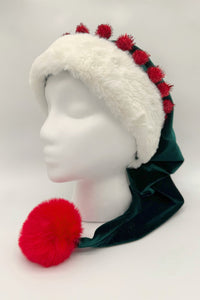 A festive elf-style hat displayed on a white mannequin head. The hat is made of dark green Velvet with a wide white faux fur trim, decorated with small red tinsel pom-poms along the top. A large fluffy red pom-pom hangs from the end of the long, pointed tail of the hat, draping down over the mannequin’s shoulder.