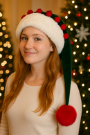 A young woman with long strawberry blonde hair smiles softly while wearing a green Velvet Santa-style hat with a wide white faux fur trim, red pom-pom accents along the brim, and an extended tail ending in a large red pom-pom that hangs about eight inches below her shoulder. She is dressed in a cream top and stands in front of a warmly lit Christmas tree decorated with lights and ornaments.