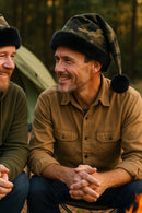 Two middle-aged men sitting together at a campsite, both wearing camouflage Santa-style hats with black faux fur trim and black pom-poms. One man is dressed in a green hoodie, the other in a tan button-up shirt, and they are smiling at each other with a tent and campfire in the background.