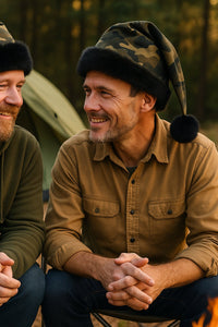 Two middle-aged men sitting together at a campsite, both wearing camouflage Santa-style hats with black faux fur trim and black pom-poms. One man is dressed in a green hoodie, the other in a tan button-up shirt, and they are smiling at each other with a tent and campfire in the background.
