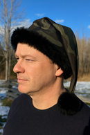 A middle-aged man outdoors on a sunny winter day wearing a camouflage Santa-style hat with black faux fur trim and a black pom-pom, standing in front of a snowy landscape with trees and a wooden fence in the background.