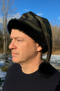 A middle-aged man outdoors on a sunny winter day wearing a camouflage Santa-style hat with black faux fur trim and a black pom-pom, standing in front of a snowy landscape with trees and a wooden fence in the background.