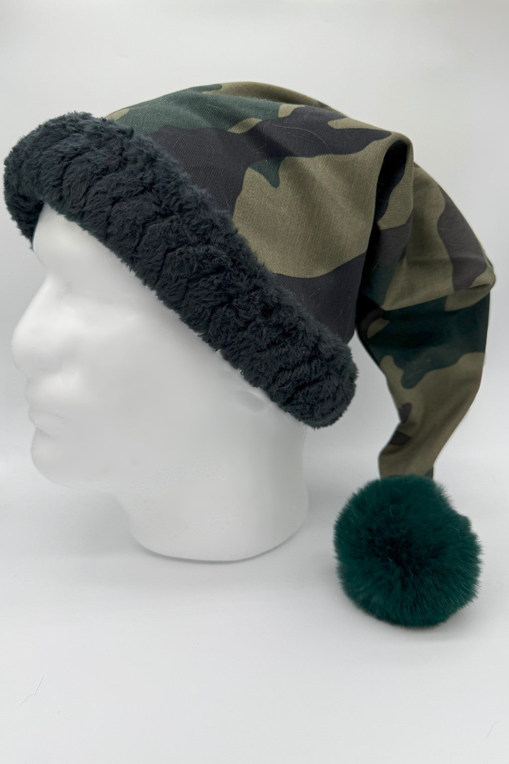 Camouflage Santa-style hat with black faux fur trim and a large forrest green fluffy pom-pom, displayed on a white mannequin head against a plain background.