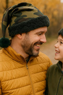 A smiling father and young son outdoors wearing matching camouflage Santa-style hats with dark faux fur trim and green pom-poms, looking at each other warmly against a backdrop of autumn trees.