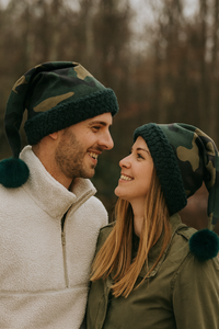 A smiling couple outdoors wearing matching camouflage Santa-style hats with dark faux fur trim and green pom-poms, standing close together while looking into each other’s eyes.