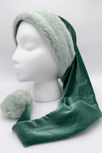 A mannequin head displays a festive Santa-style hat with a wide, soft faux fur trim in light mint green. The body of the hat is made from shimmering mossy green Velvet with a subtle diamond pattern, ending in a large fluffy mint green pom-pom that rests on the surface.