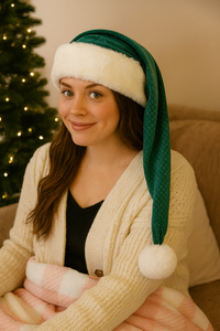 A young woman with long brown hair is smiling softly while wearing a milky mint–colored Santa hat with a white faux fur trim and fluffy pom-pom. She is dressed in a cream sweater and sits indoors in front of a warmly lit Christmas tree with twinkling lights, creating a cozy holiday atmosphere.