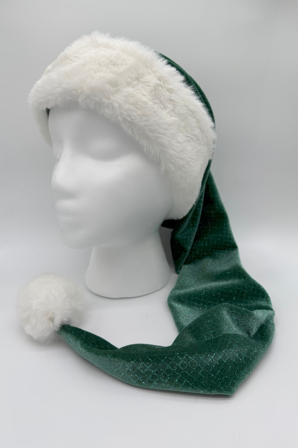A long green Velvet Santa-style hat with a white faux fur trim and fluffy white pom-pom, displayed on a white mannequin head against a plain background.