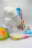 long Santa-style hat displayed on a white mannequin head. The hat features a soft white faux fur trim around the face opening, a pastel tie-dye fabric in blue, pink, yellow, and orange along the length, and ends with a large fluffy pom-pom in vibrant rainbow colors of yellow, orange, green, and blue.