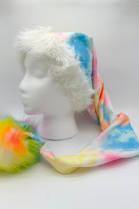 long Santa-style hat displayed on a white mannequin head. The hat features a soft white faux fur trim around the face opening, a pastel tie-dye fabric in blue, pink, yellow, and orange along the length, and ends with a large fluffy pom-pom in vibrant rainbow colors of yellow, orange, green, and blue.