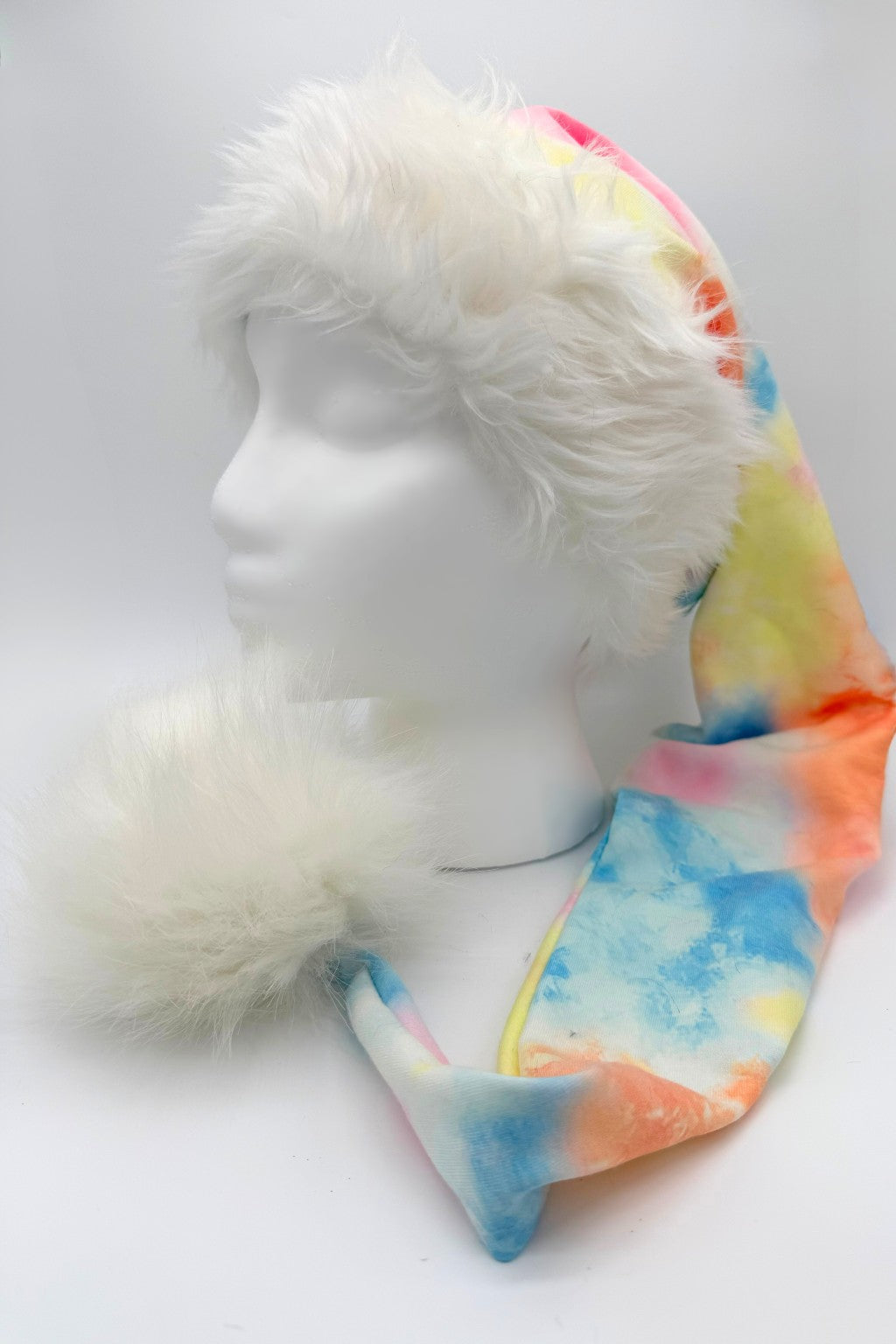 A colorful Santa-style hat with a fluffy white faux fur brim and large white pom-pom, displayed on a white mannequin head. The body of the hat features a tie-dye pattern in blue, orange, yellow, and pink shades.
