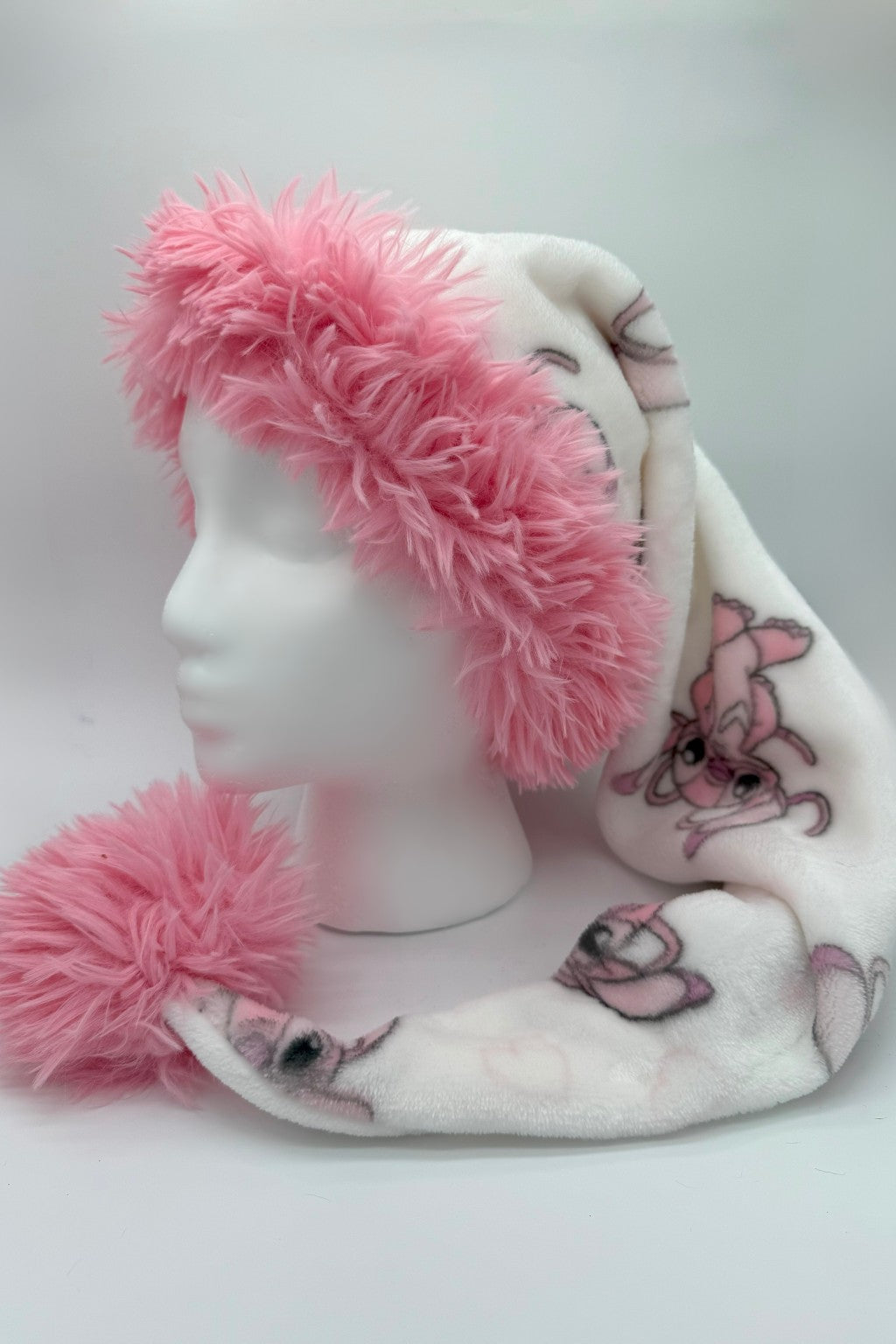 A white fleece Santa-style hat with light pink cartoon character of Angel from the Disney movie Stitch, trimmed in fluffy pink faux fur, and finished with a large pink faux fur pom-pom at the tip. The hat is displayed on a white mannequin head against a neutral background.