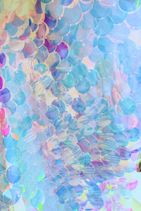 A close-up of iridescent, overlapping sequins in shades of Teal, blue, purple, and yellow, attached to a sheer mesh fabric. The sequins catch the light, creating a shimmering, rainbow-like effect with a textured, layered surface.