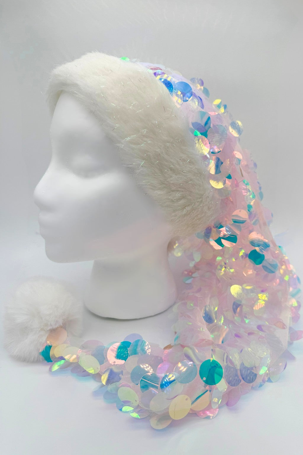 A festive Santa-style hat displayed on a white mannequin head. The hat has a fluffy white faux fur trim and a matching white pom-pom at the end. The body of the hat is covered in sheer fabric with large iridescent sequins that shimmer in pastel shades of pink, blue, green, and yellow, creating a sparkling, mermaid-like effect.