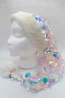 A festive Santa-style hat displayed on a white mannequin head. The hat has a fluffy white faux fur trim and a matching white pom-pom at the end. The body of the hat is covered in sheer fabric with large iridescent sequins that shimmer in pastel shades of pink, blue, green, and yellow, creating a sparkling, mermaid-like effect.