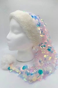 A festive Santa-style hat displayed on a white mannequin head. The hat has a fluffy white faux fur trim and a matching white pom-pom at the end. The body of the hat is covered in sheer fabric with large iridescent sequins that shimmer in pastel shades of pink, blue, green, and yellow, creating a sparkling, mermaid-like effect.