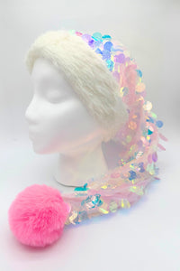 A festive Santa-style hat displayed on a white mannequin head. The hat has a fluffy white faux fur trim and a long, draping body made of sheer pink fabric covered in iridescent sequins that reflect pastel shades of blue, purple, and green. At the end of the hat hangs a large, round, bright pink faux fur pom-pom.