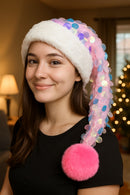 A young woman with long brown hair smiles softly indoors while wearing a festive Santa-style hat. The hat features a fluffy white faux fur brim, an iridescent pastel sequin-covered tail, and a large bright pink faux fur pom-pom that hangs down over her shoulder. She is dressed in a simple black top, with warm indoor lighting, a lamp, and a decorated Christmas tree visible in the background.