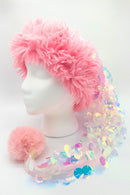 A festive hat displayed on a white mannequin head. The hat features a fluffy pink faux-fur band and an elongated tail covered in iridescent sequins that shimmer with pastel colors. At the end of the tail is a large, soft pink pom-pom resting on the surface. The sequins reflect rainbow hues, giving the hat a whimsical and playful appearance.