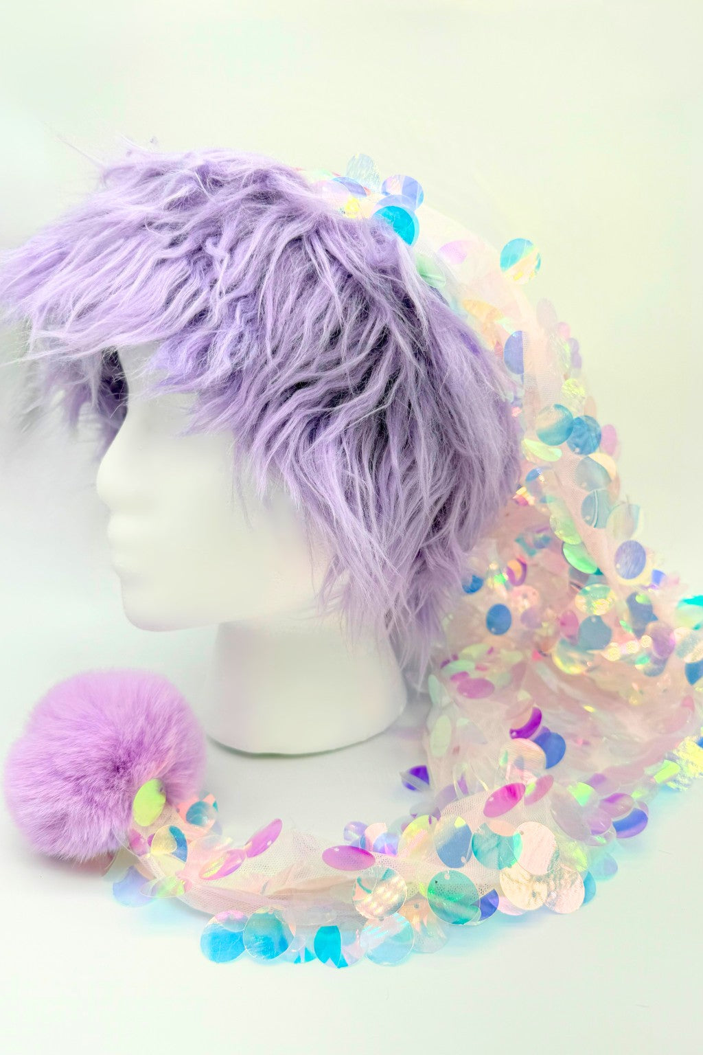 A mannequin head with shaggy lavender faux-fur hair wearing a whimsical pastel hat. The hat features an iridescent sequin-covered fabric that drapes down, finished with a fluffy lavender pom-pom at the end. The sequins reflect rainbow hues of pink, blue, green, and gold, giving the hat a magical, mermaid-like appearance.