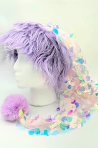 A mannequin head with shaggy lavender faux-fur hair wearing a whimsical pastel hat. The hat features an iridescent sequin-covered fabric that drapes down, finished with a fluffy lavender pom-pom at the end. The sequins reflect rainbow hues of pink, blue, green, and gold, giving the hat a magical, mermaid-like appearance.