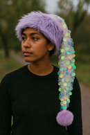 A young woman with medium brown skin wears a lavender faux fur Santa-style hat with a long iridescent sequin-covered tail and a fluffy lavender pom-pom at the end, standing outdoors with blurred greenery in the background.