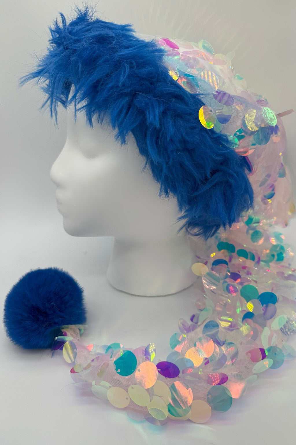 A Santa-style hat with bright blue faux fur trim, a long tail of iridescent pastel sequins, and a fluffy blue pom-pom displayed on a white mannequin head.