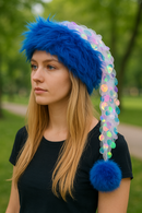 A young woman with long blond hair is wearing a vibrant blue faux fur hat with a cascading tail of sheer fabric decorated with large iridescent sequins, ending in a fluffy blue pom-pom. She is outdoors in a park setting, dressed in a simple black top, with trees and a pathway blurred in the background.