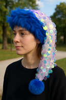 A young woman with fair skin and short brown hair is wearing a vibrant blue faux fur hat with a long tail of sheer fabric adorned with large iridescent sequins, ending in a fluffy blue pom-pom. She is outdoors in a park, dressed in a plain black top, with trees and a walking path blurred in the background.