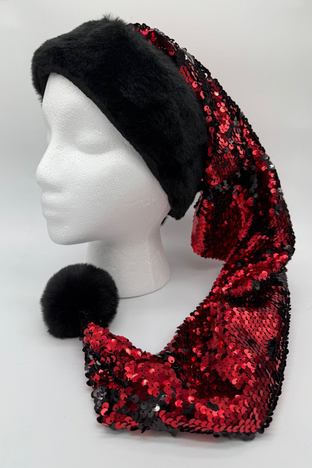 A mannequin head wearing a festive Santa-style hat with a black faux fur brim and a long, pointed body covered in shiny red and black sequins, ending in a black faux fur pom-pom.