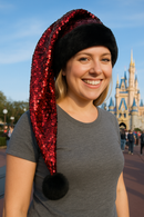 A smiling young woman at an amusement park wears a festive Santa-style hat with a black faux fur brim, a long red sequin tail, and a fluffy black pom-pom that hangs about eight inches below her shoulder. She is the main focus of the photo, with the blurred outline of a Castle and other visitors in the background under a clear blue sky.